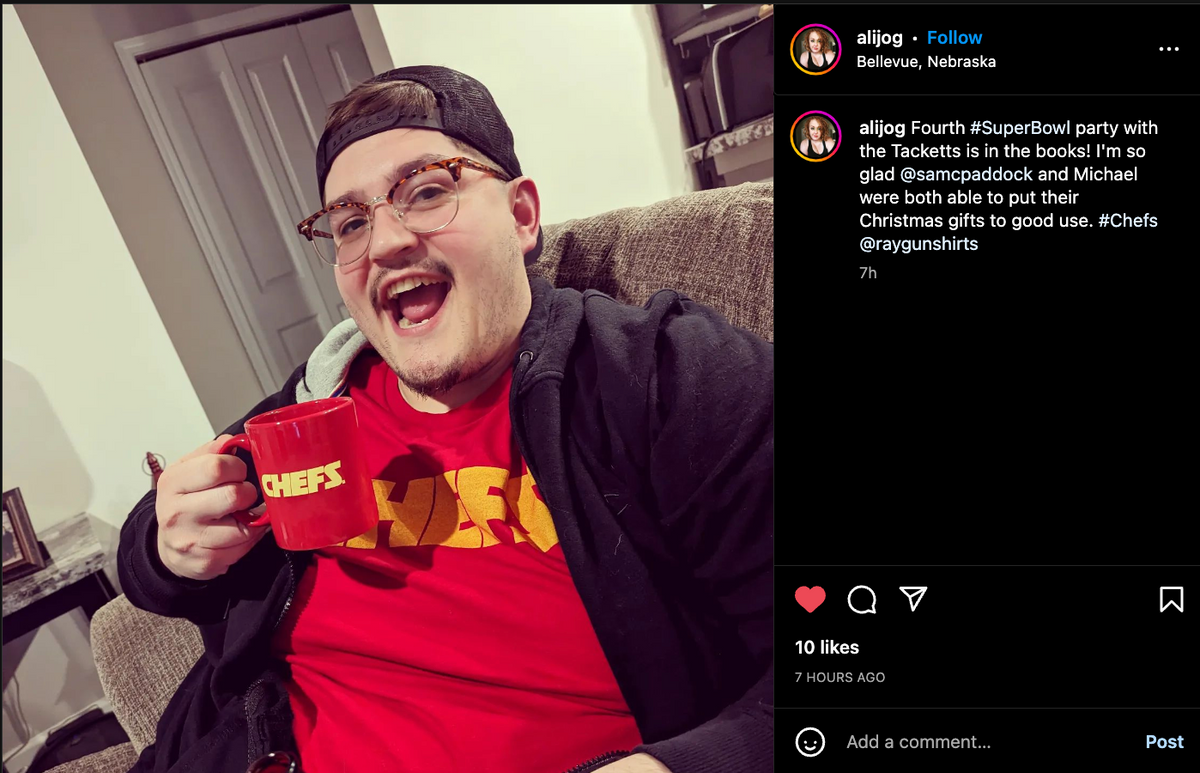 A smiling man wearing a gold screen-print Kansas City Chiefs t-shirt and a baseball cap holds a red CHEFS mug.