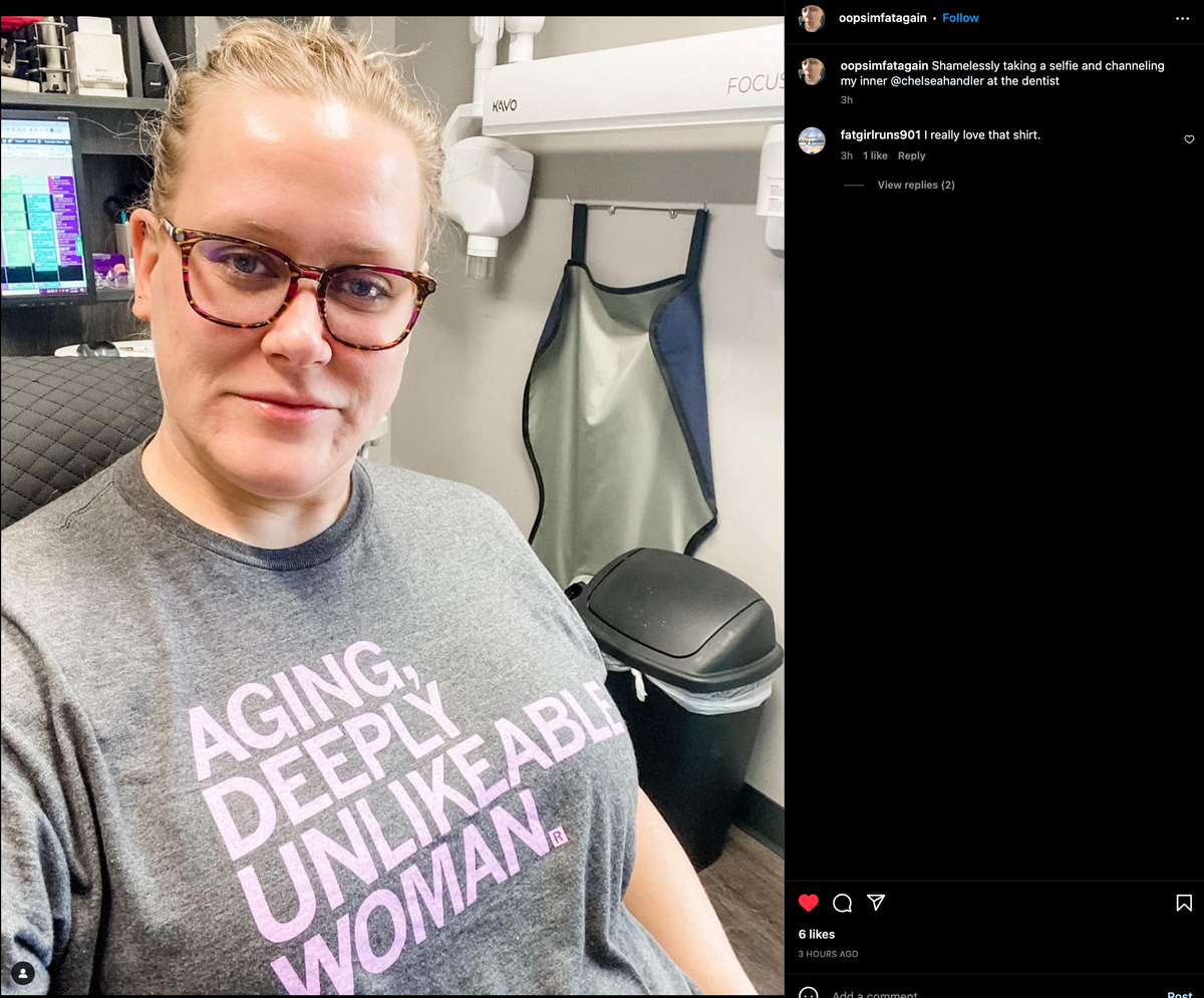 A woman wearing glasses and a charcoal T-shirt with the text AGING, DEEPLY UNLIKEABLE WOMAN in lilac screen print, taking a selfie in a dentist’s office.