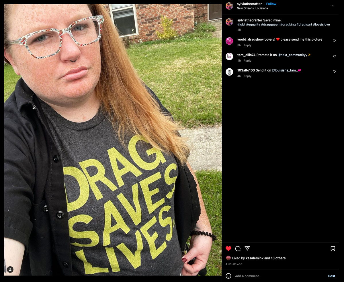 A person with red hair and glasses wearing a charcoal t-shirt that says DRAG SAVES LIVES in yellow lettering.