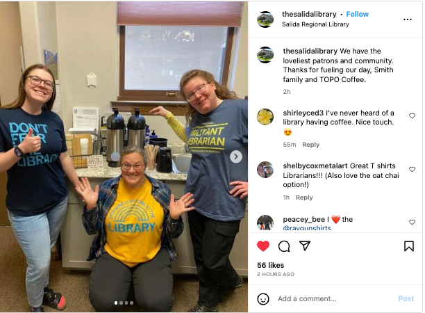 Three smiling librarians wearing a library t-shirt, a Militant Librarian shirt, and an Everyone is Welcome at the Library shirt, standing in a breakroom near a coffee station.