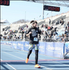 A long-haired runner wearing a black shirt with the text Call for help: Theres hope crosses the finish line at a track stadium, supporting the 988 lifeline.