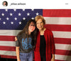A young woman wearing a union made shirt that reads America Needs Female Leaders poses with Elizabeth Warren in front of an American flag.
