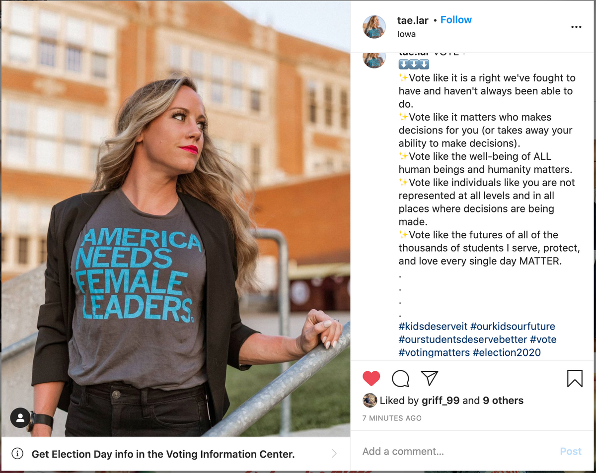 A woman wears a union made, grey t-shirt that says AMERICA NEEDS FEMALE LEADERS in blue lettering while standing outdoors in front of a school building.
