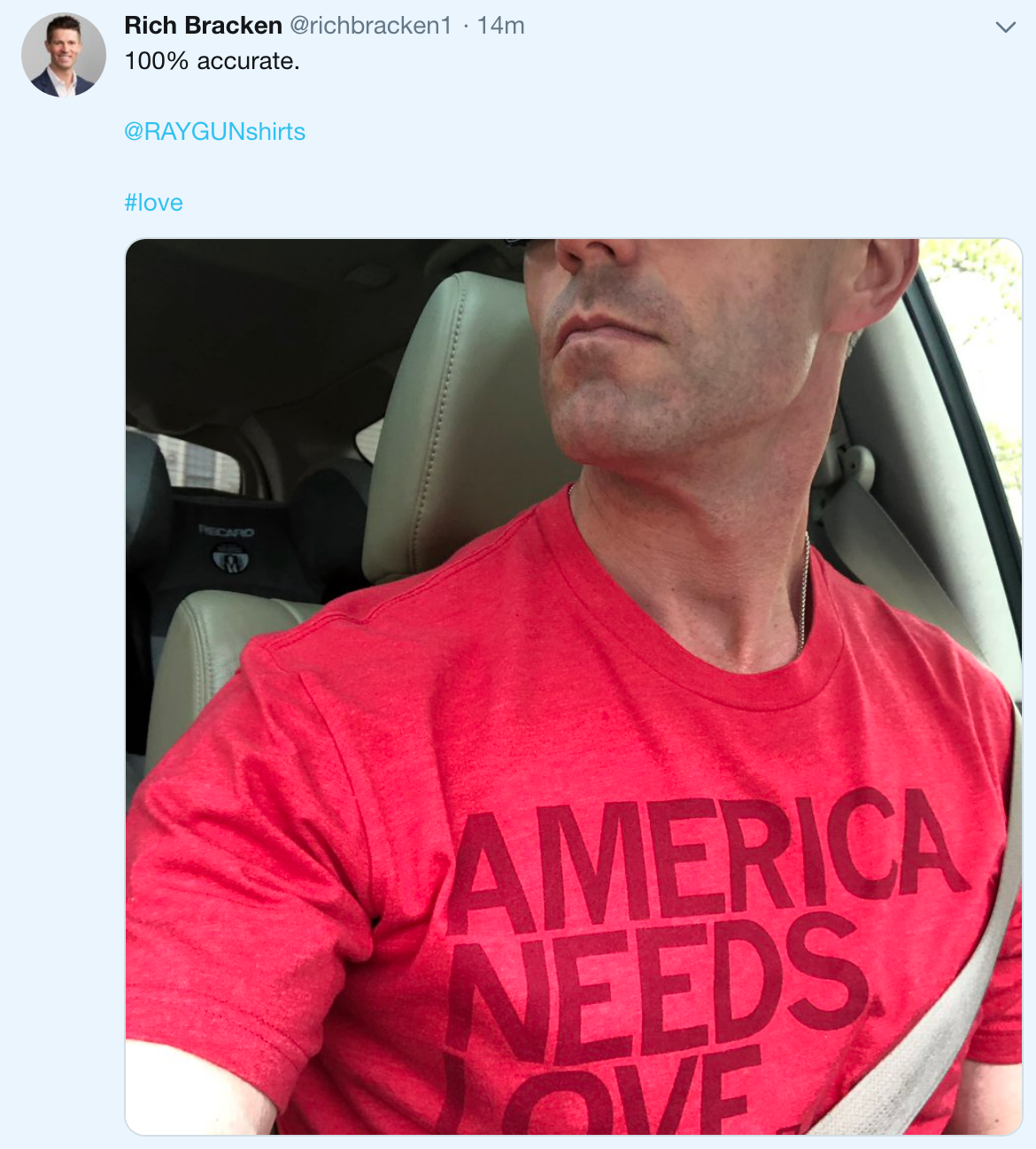 A man sits in a car wearing a heather red t-shirt that says AMERICA NEEDS LOVE.