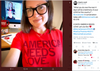 A woman wearing black glasses and a heather red T-shirt that says AMERICA NEEDS LOVE smiles at the camera, with a computer monitor showing a video call in the background.