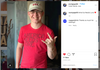 A person wearing a USA baseball cap and a heather red t-shirt that says America Needs Love makes a hand gesture.