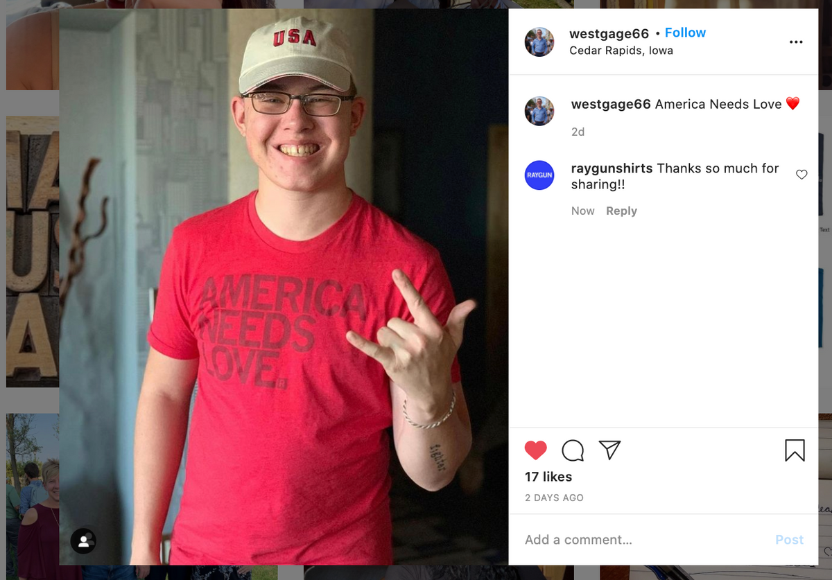 A person wearing a USA baseball cap and a heather red t-shirt that says America Needs Love makes a hand gesture.