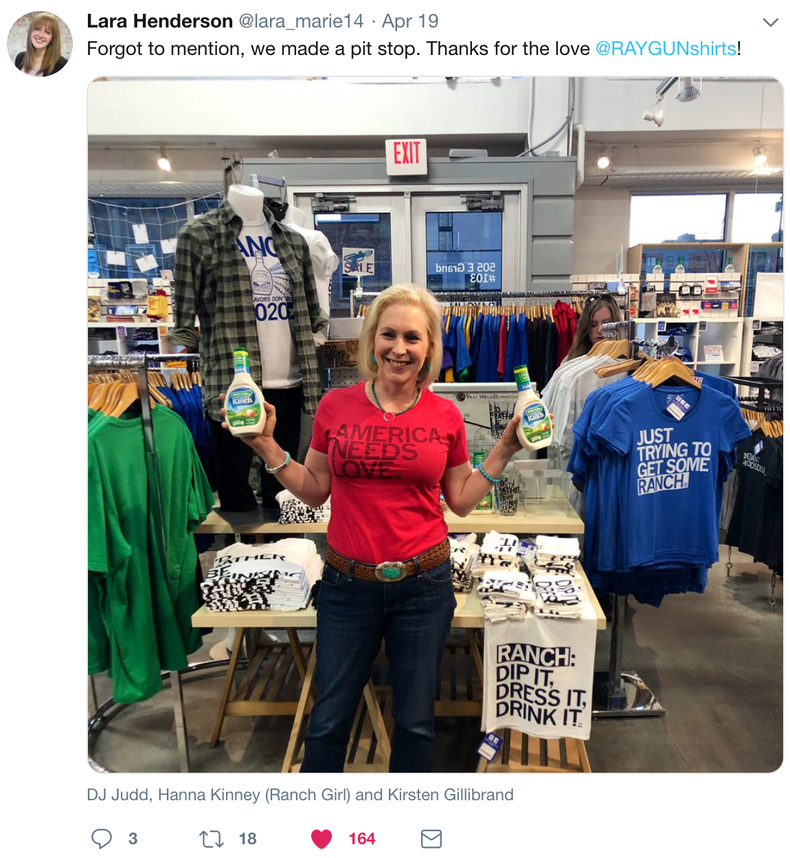 Kirsten Gillibrand stands in a store holding two bottles of ranch dressing, wearing a heather red t-shirt that reads America Needs Love above racks of clothing.