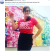 A smiling woman wearing glasses and a union-made pink t-shirt that says America needs public schools holds an apple in front of a colorful student art display.