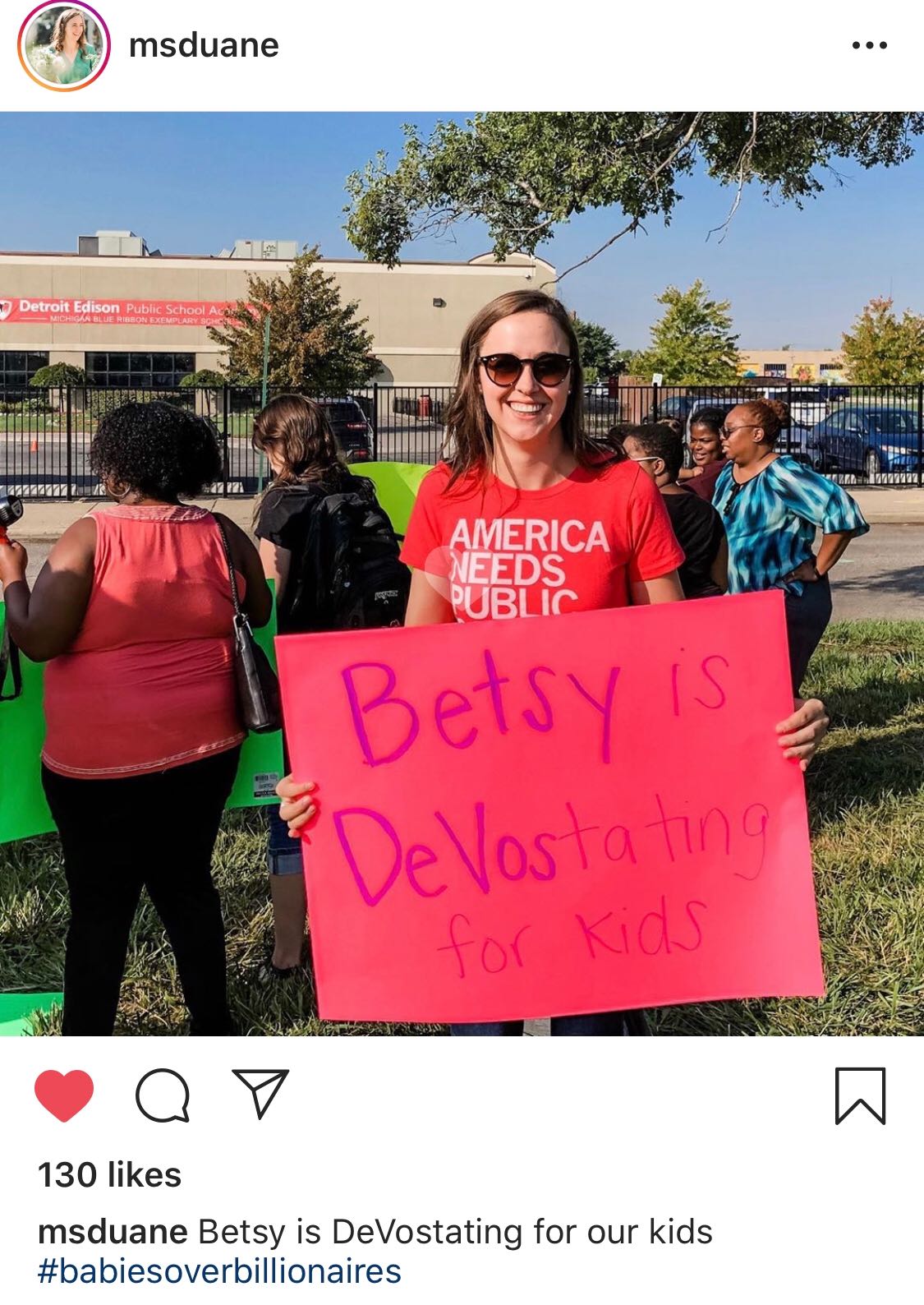 A woman wearing a union made America Needs Public t-shirt holds a pink protest sign that says Betsy is DeVostating for Kids in front of a public school.