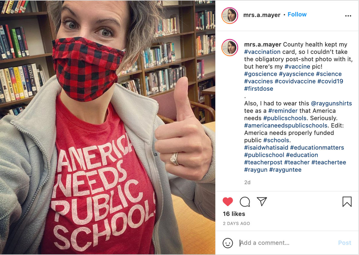 A woman wearing a red and black buffalo plaid face mask and a union made t-shirt that reads America needs public schools gives a thumbs-up in a library.