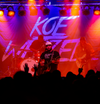 Koe Wetzel performs on stage in a union made shirt in front of a large backdrop featuring his name, illuminated by red and orange concert lighting.