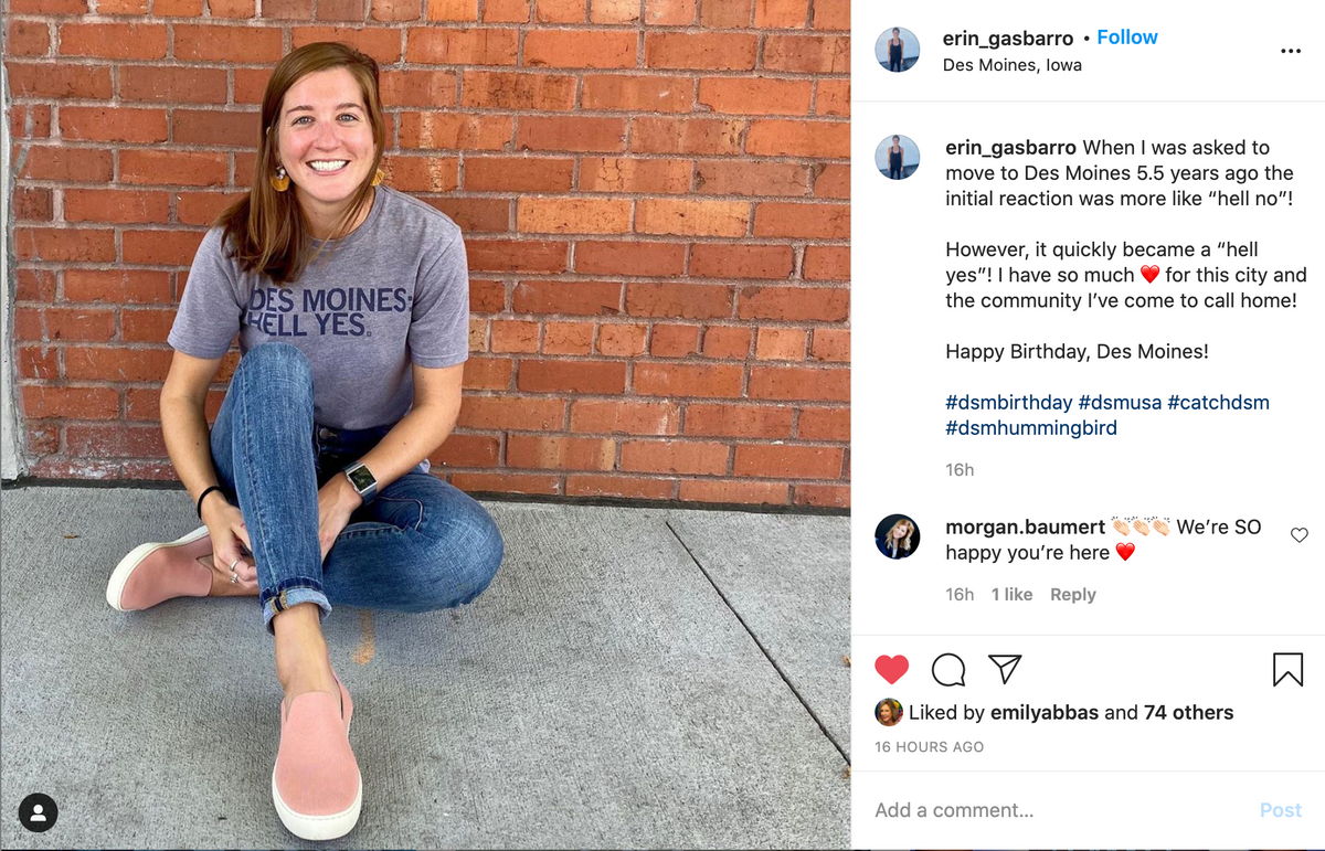 A smiling woman sits on the ground against a red brick wall, wearing a heather grey T-shirt that says DES MOINES HELL YES and pink slip-on sneakers.