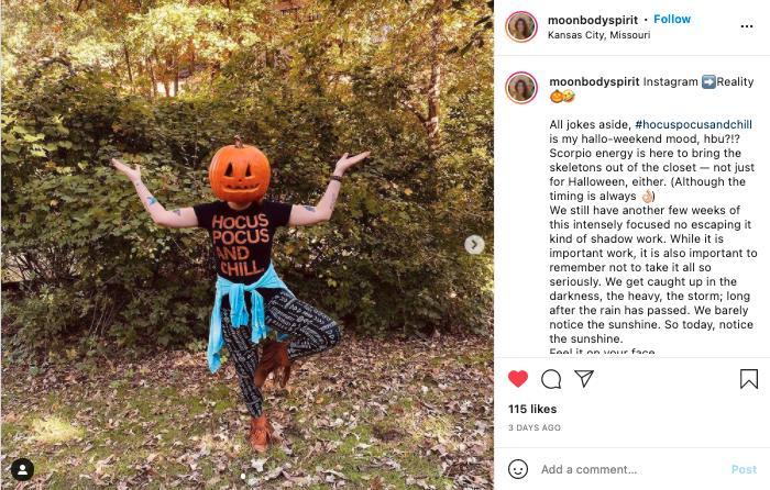 A person with a pumpkin head performs a tree yoga pose in a park, wearing a black Hocus Pocus and Chill t-shirt.