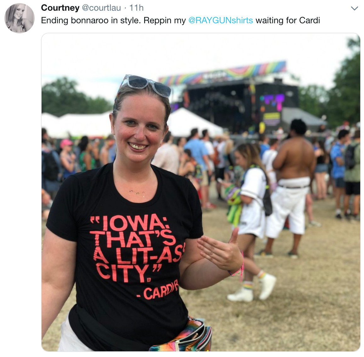 A smiling woman stands at a music festival wearing a black t-shirt featuring a retired Cardi B design that reads: IOWA: THATS A LIT-AS CITY.