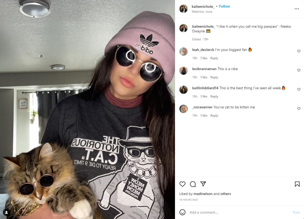 A woman in a pink beanie and circular sunglasses holds a Notorious CAT wearing matching circular sunglasses and a gold chain, pictured on a charcoal T-shirt.