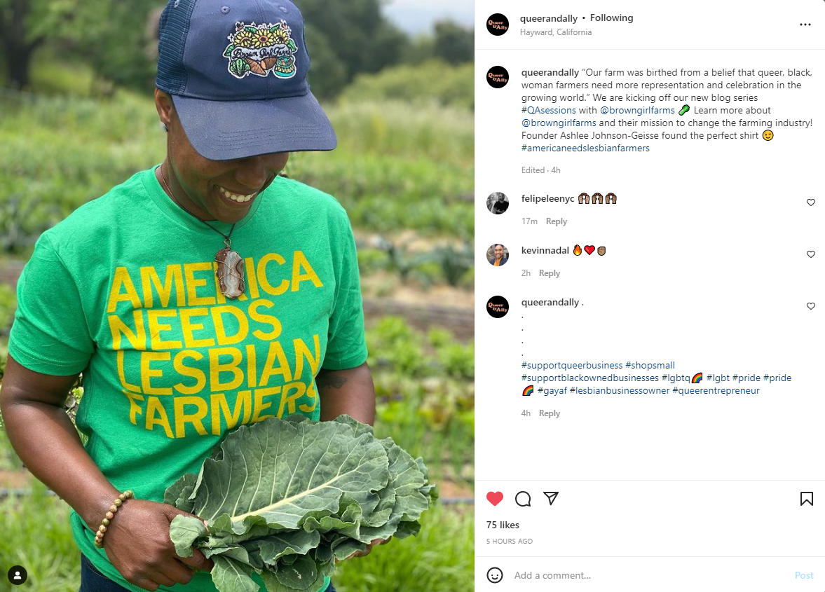 A smiling person in a baseball cap and a heather kelly green t-shirt that reads America Needs Lesbian Farmers holds a large bunch of kale in a field.