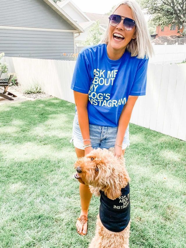 A woman laughing in a yard while wearing a blue Instagram dog shirt, standing with a dog wearing a matching black shirt from RAYGUN dog apparel.