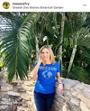 A woman wears an eco-friendly blue t-shirt featuring a globe graphic and the text Keep It Clean: This Is Earth, Not Uranus while standing in front of palm trees.