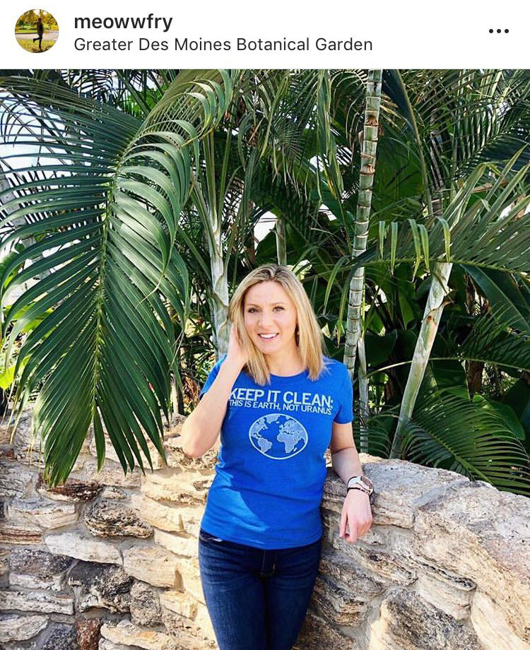 A woman wears an eco-friendly blue t-shirt featuring a globe graphic and the text Keep It Clean: This Is Earth, Not Uranus while standing in front of palm trees.
