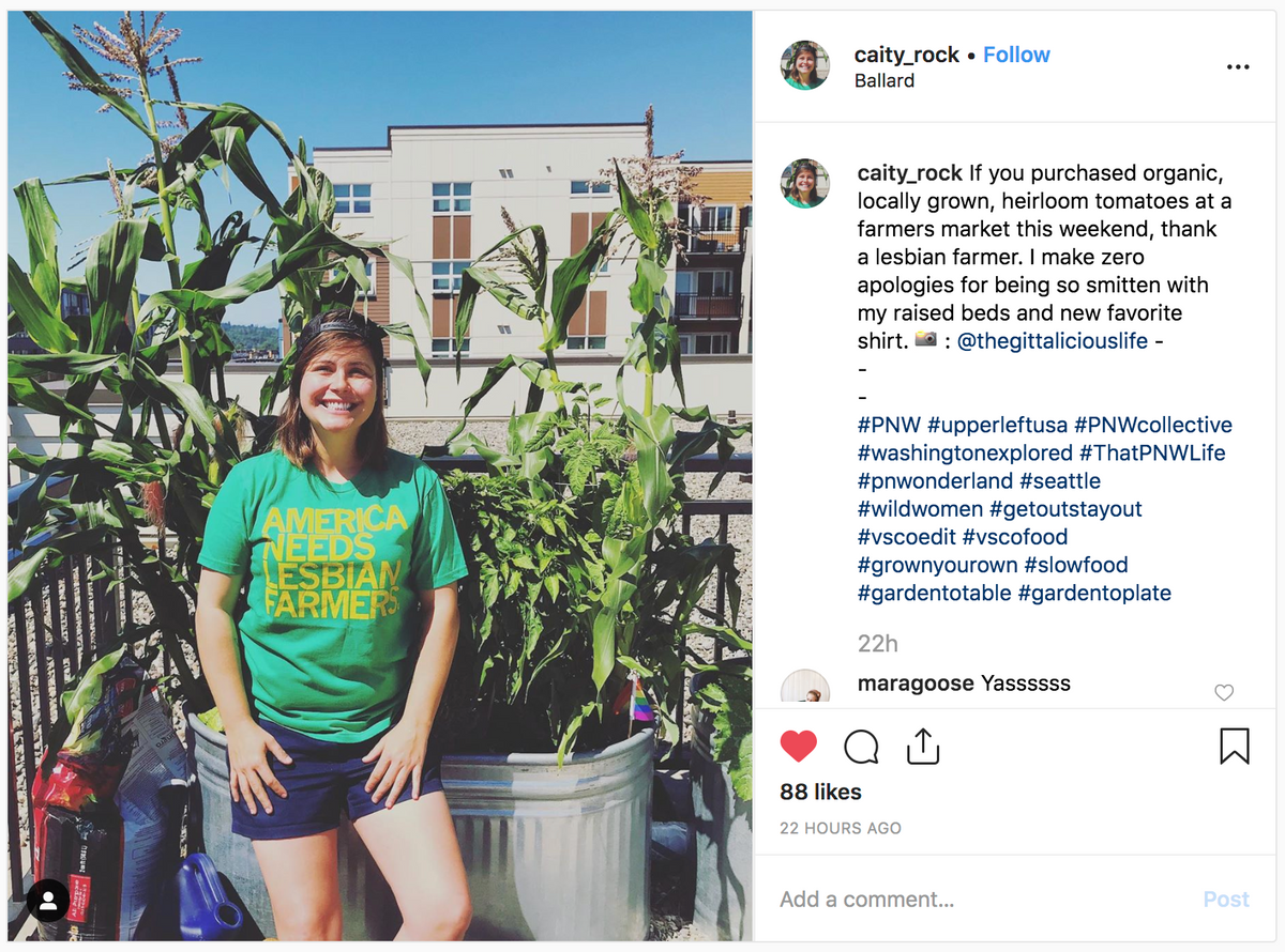 A woman stands in a garden wearing a heather kelly green t-shirt that says America Needs Lesbian Farmers in front of tall corn plants and a metal planter.