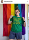 A person wearing glasses and a heather kelly green T-shirt that reads AMERICA NEEDS LESBIAN FARMERS stands in front of a rainbow pride flag.
