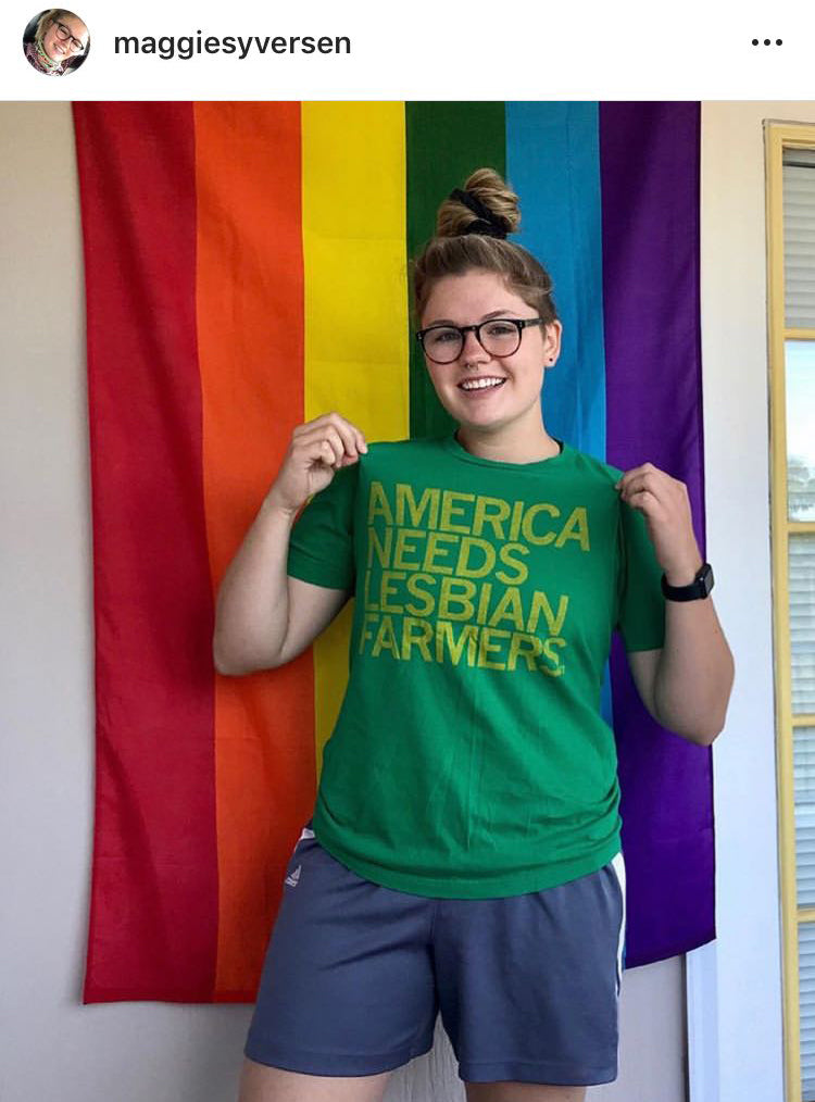 A person wearing glasses and a heather kelly green T-shirt that reads AMERICA NEEDS LESBIAN FARMERS stands in front of a rainbow pride flag.