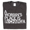 A folded heather black shirt with all caps white text that says, "A Woman's Place Is In Charge"