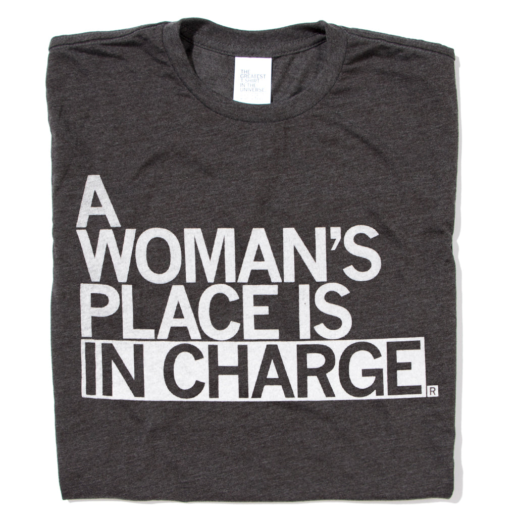 A folded heather black shirt with all caps white text that says, "A Woman's Place Is In Charge"