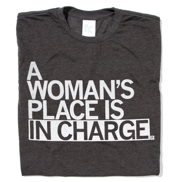 A folded heather black shirt with all caps white text that says, "A Woman's Place Is In Charge"