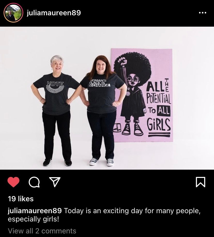 Two women stand next to a large graphic of a girl with an afro raising her fist, captioned The potential to all girls. They wear union made shirts reading Vote and A woman’s place is in charge, promoting women’s empowerment.